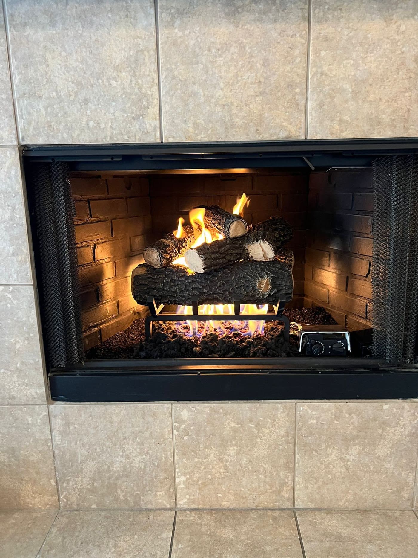 Vented gas log set running in a masonry firebox with a tiled surround, the decorative flames orange over the logs and the burner line beneath them burning blue