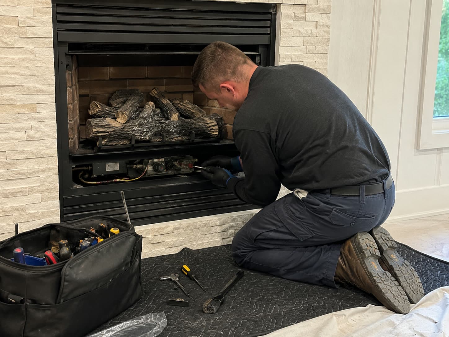 Chimney technician kneeling on a protective mat servicing the valve assembly of a gas fireplace, with the lower access panel removed and hand tools laid out beside an open tool bag